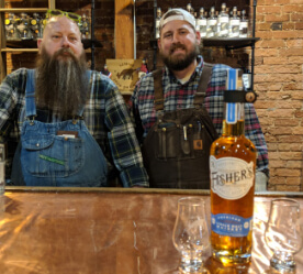 Two bearded men in plaid and overalls with a bottle of whiskey