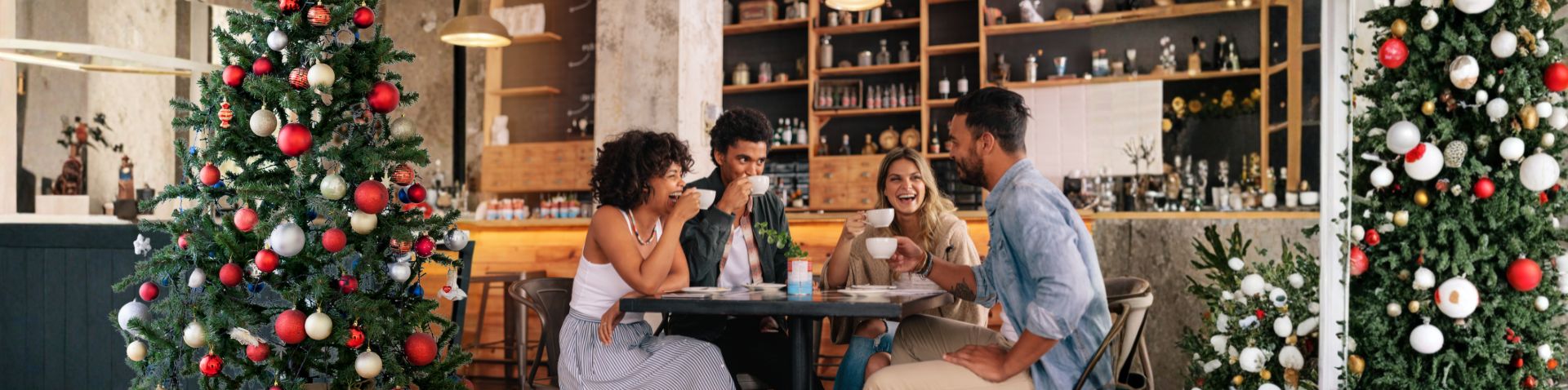A group of four friends enjoying coffee and laughing around a table in a stylish cafe decorated with large Christmas trees.